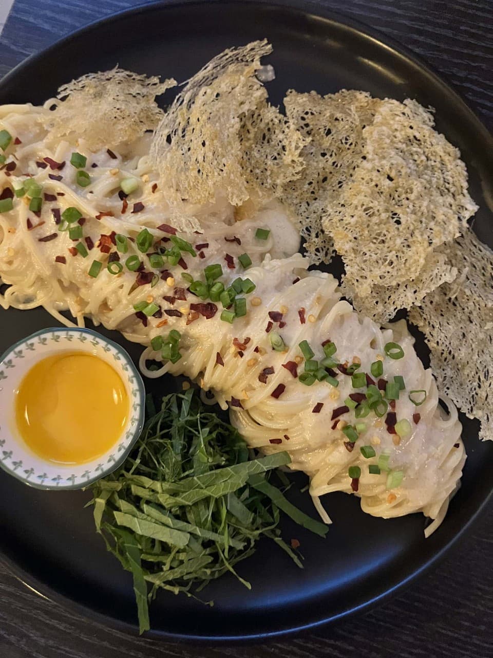 어두운 나무 테이블 위 검은 접시에 담긴 크림 면 요리와 노른자 소스, 채 썬 깻잎과 얇은 그물 형태의 바삭한 튀김