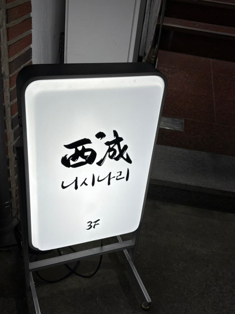 어두운 벽돌 건물 입구에 세워진 흰색 사각형 조명 입간판과 검은색 붓글씨로 적힌 층수 안내 문구