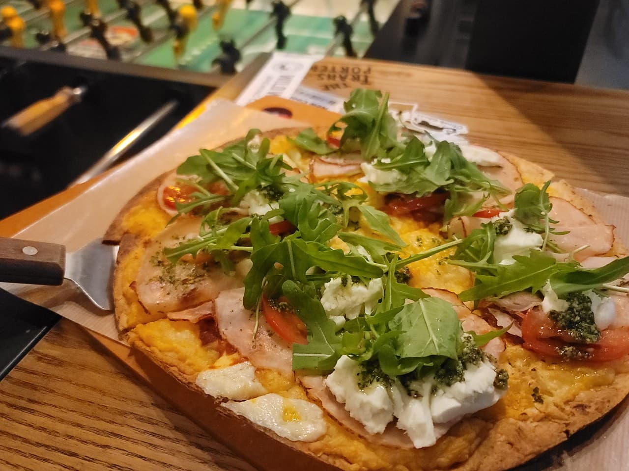 목재 바 카운터 위에 놓인 루꼴라와 치즈가 토핑된 피자와 배경에 보이는 테이블 축구 게임기