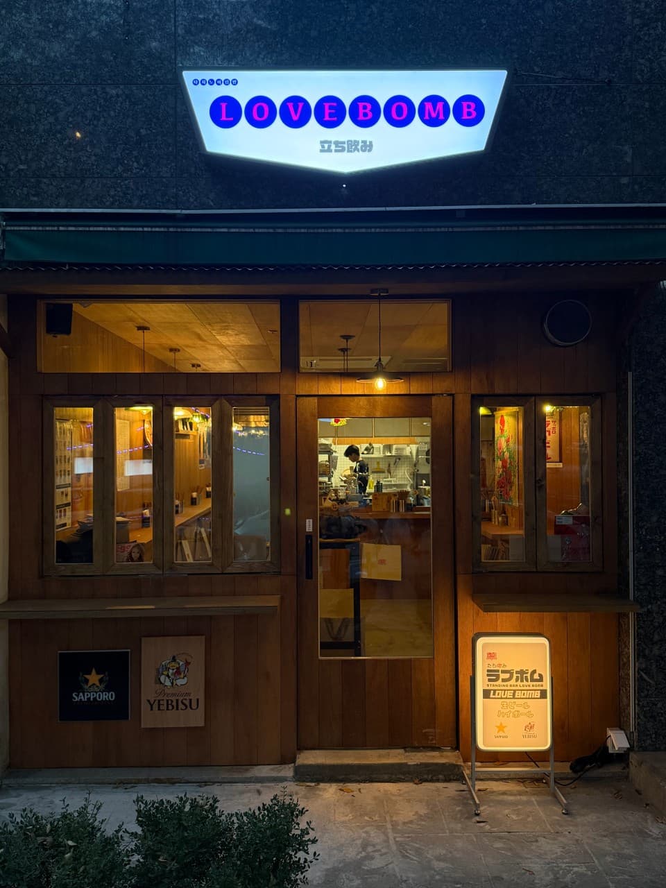 어두운 목재 외관과 유리창 너머 주황색 조명이 비치는 1인용 나무 카운터석. 상단 흰색 간판과 입간판이 켜진 저녁 무렵의 입구