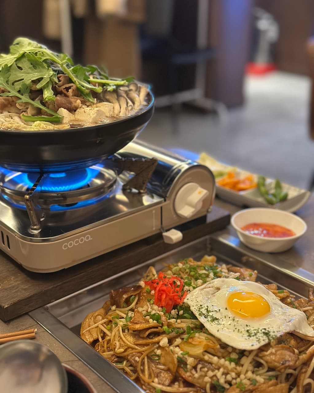 파란 불꽃의 휴대용 버너 위 소고기 전골과 계란 프라이를 얹은 볶음면이 차려진 1인용 테이블