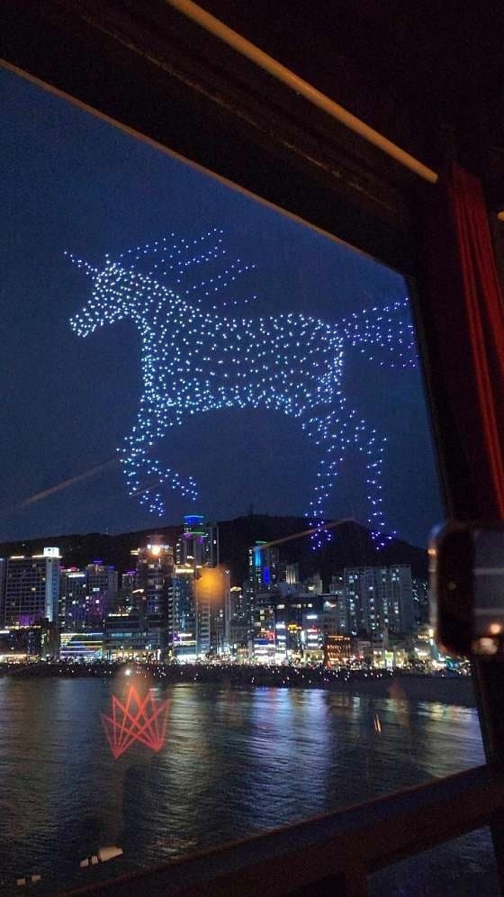 어두운 실내 창가 좌석에서 내다본 밤하늘의 유니콘 형상 드론 쇼와 불 켜진 고층 빌딩이 늘어선 해변 전경