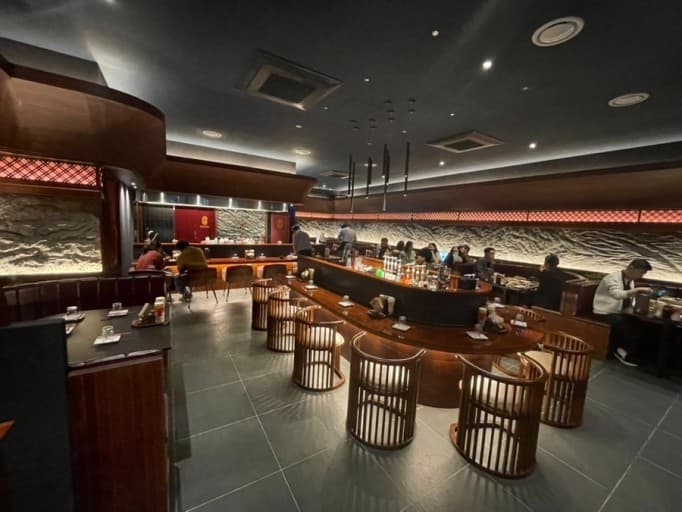 오렌지색 조명이 비치는 돌 벽면과 중앙의 긴 곡선형 나무 카운터석, 1인용 목재 의자들이 배치된 공간