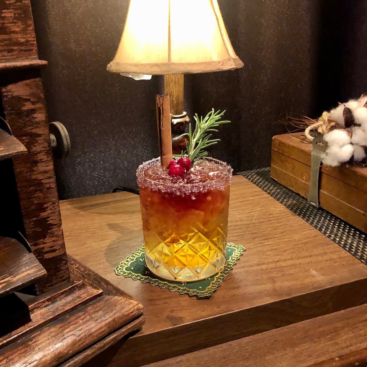 황색 조명 아래 나무 테이블 위 시나몬 스틱과 로즈마리가 꽂힌 칵테일 잔과 녹색 코스터