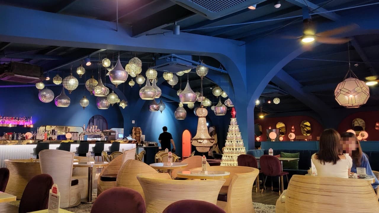 파란색 천장 아래 매달린 여러 개의 구형 전등과 샹들리에. 라탄 소재의 1인용 원형 의자와 소형 원형 테이블이 놓인 실내 공간