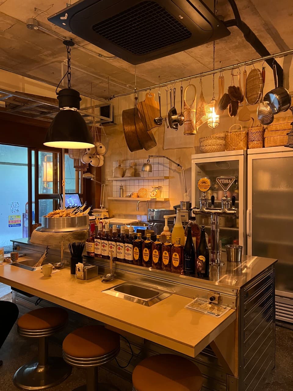 주황색 조명이 비치는 1인용 의자가 놓인 나무 카운터와 일렬로 늘어선 위스키 병, 한쪽에 놓인 오뎅 냄비와 천장에 매달린 주방 도구