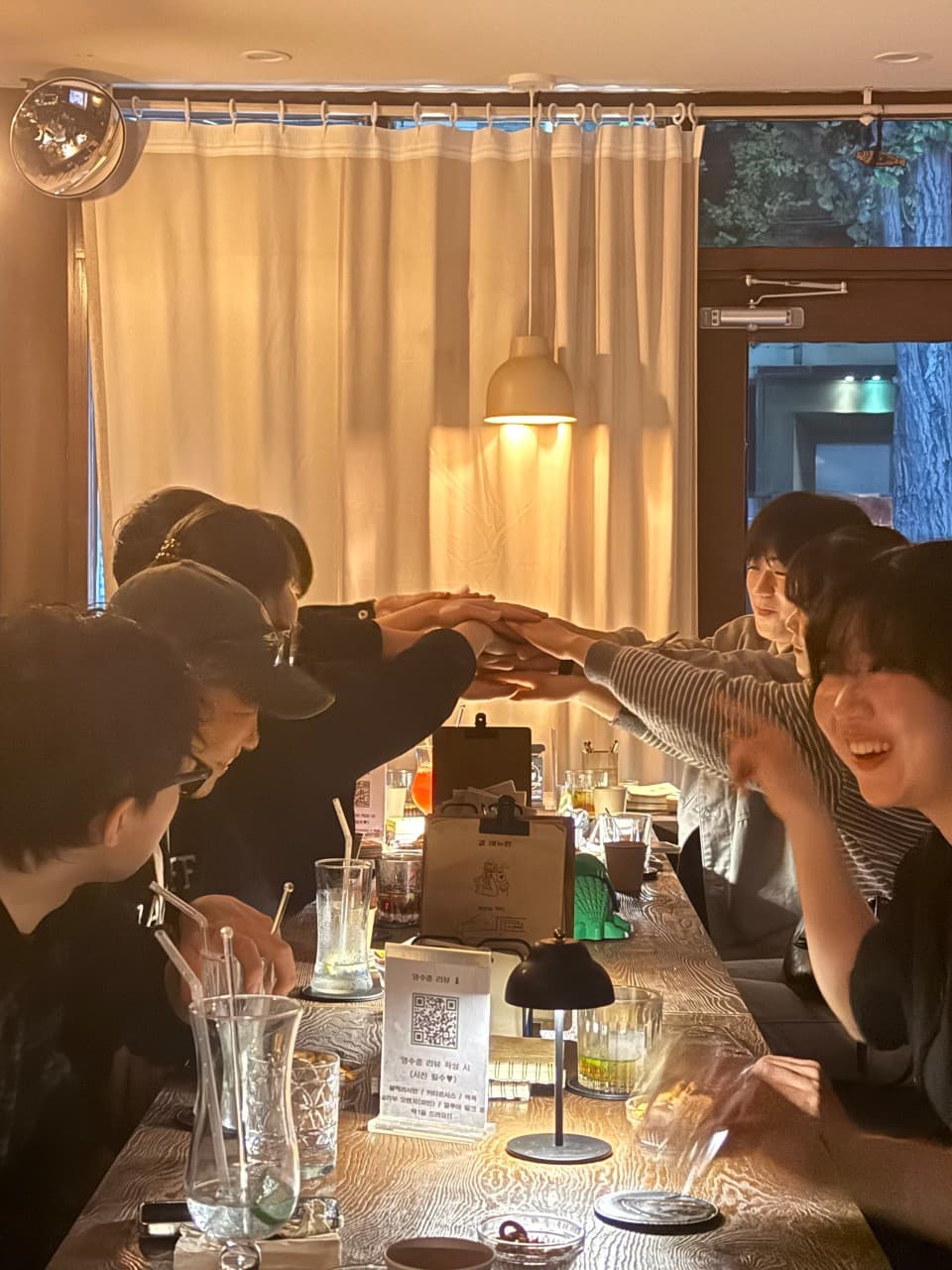노란 조명 아래 나무 카운터 석에 앉은 사람들이 중앙으로 손을 모으고 있으며 테이블 위에는 하이볼 잔과 메뉴판이 놓여 있음