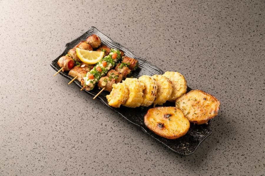 회색 석재 질감 테이블 위 검은색 긴 접시에 담긴 모둠 꼬치와 레몬 조각, 구운 옥수수와 감자 구이