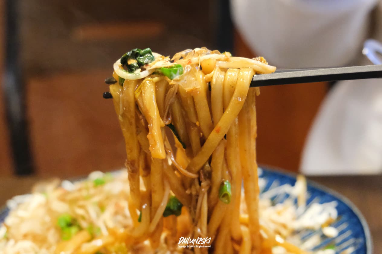 검은색 젓가락으로 들어 올린 갈색 소스의 볶음면과 대파, 숙주나물, 가쓰오부시가 곁들여진 야키소바