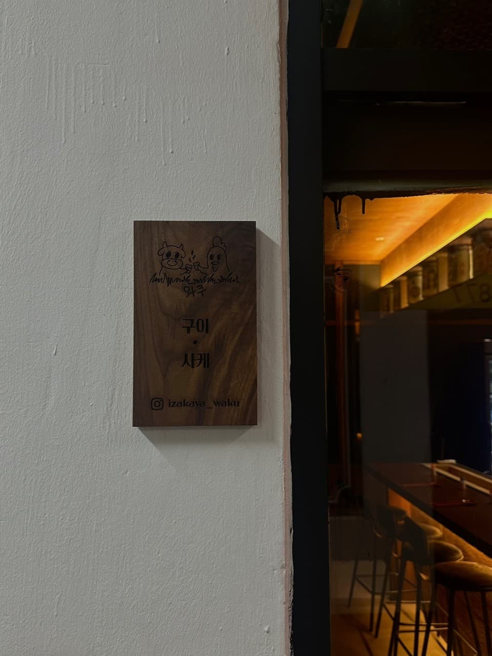 흰색 벽면의 목재 현판과 유리창 너머 주황색 조명 아래 배치된 긴 카운터석과 높은 의자