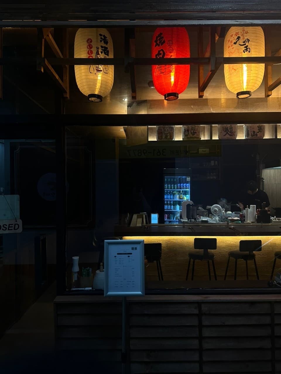 붉은색과 흰색 제등이 걸린 입구와 통유리 너머 노란 조명이 켜진 바 테이블 및 1인용 좌석