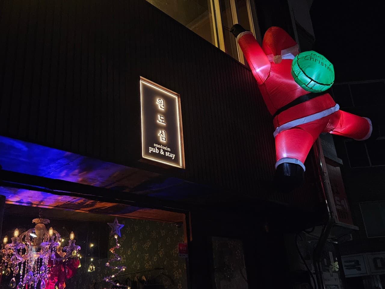 어두운 외벽에 매달린 산타 인형과 세로형 간판, 유리창 안쪽 보라색 샹들리에와 꽃무늬 벽지가 보이는 바 외관