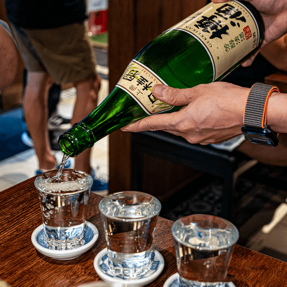 짙은 색 나무 탁자 위 작은 유리잔에 커다란 초록색 사케 병을 기울여 투명한 액체를 채우는 손길