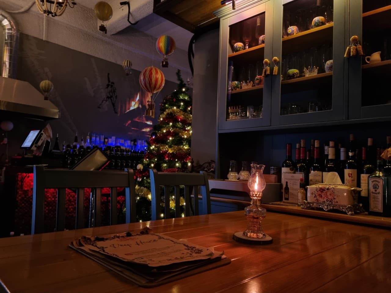 어두운 조명 아래 목재 테이블 위 메뉴판과 호롱불, 뒤편의 와인 진열장과 조명이 켜진 트리 및 공중에 매달린 열기구 장식