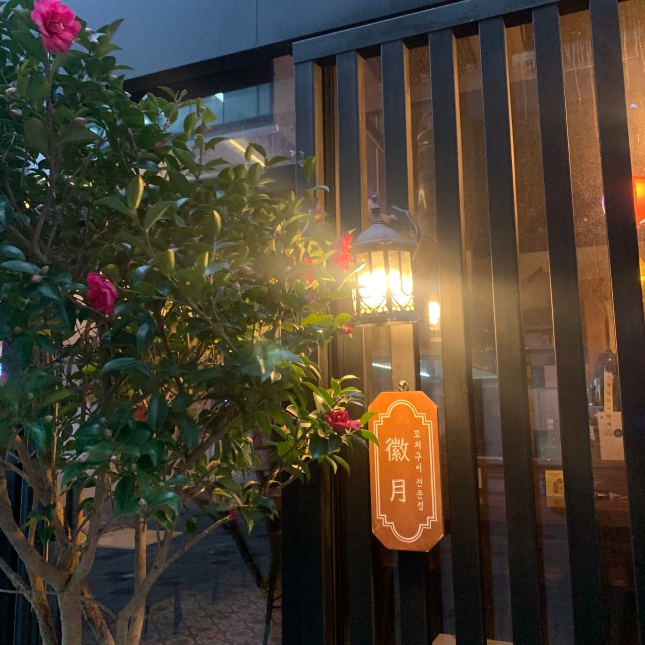 붉은 꽃나무 옆 검은색 세로 창살로 된 외벽과 황색 조명 아래 꼬치구이 전문점 문구가 적힌 작은 목재 현판