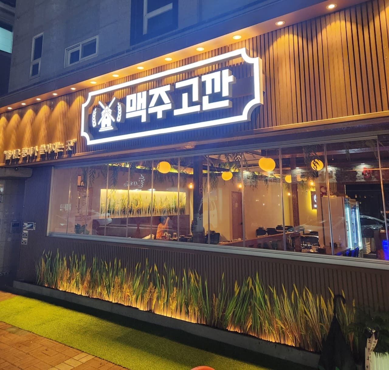 밝게 빛나는 흰색 간판과 목재 외벽의 건물 전경. 통유리창 너머로 노란 원형 조명이 켜진 바 테이블과 1인용 좌석이 배치된 내부