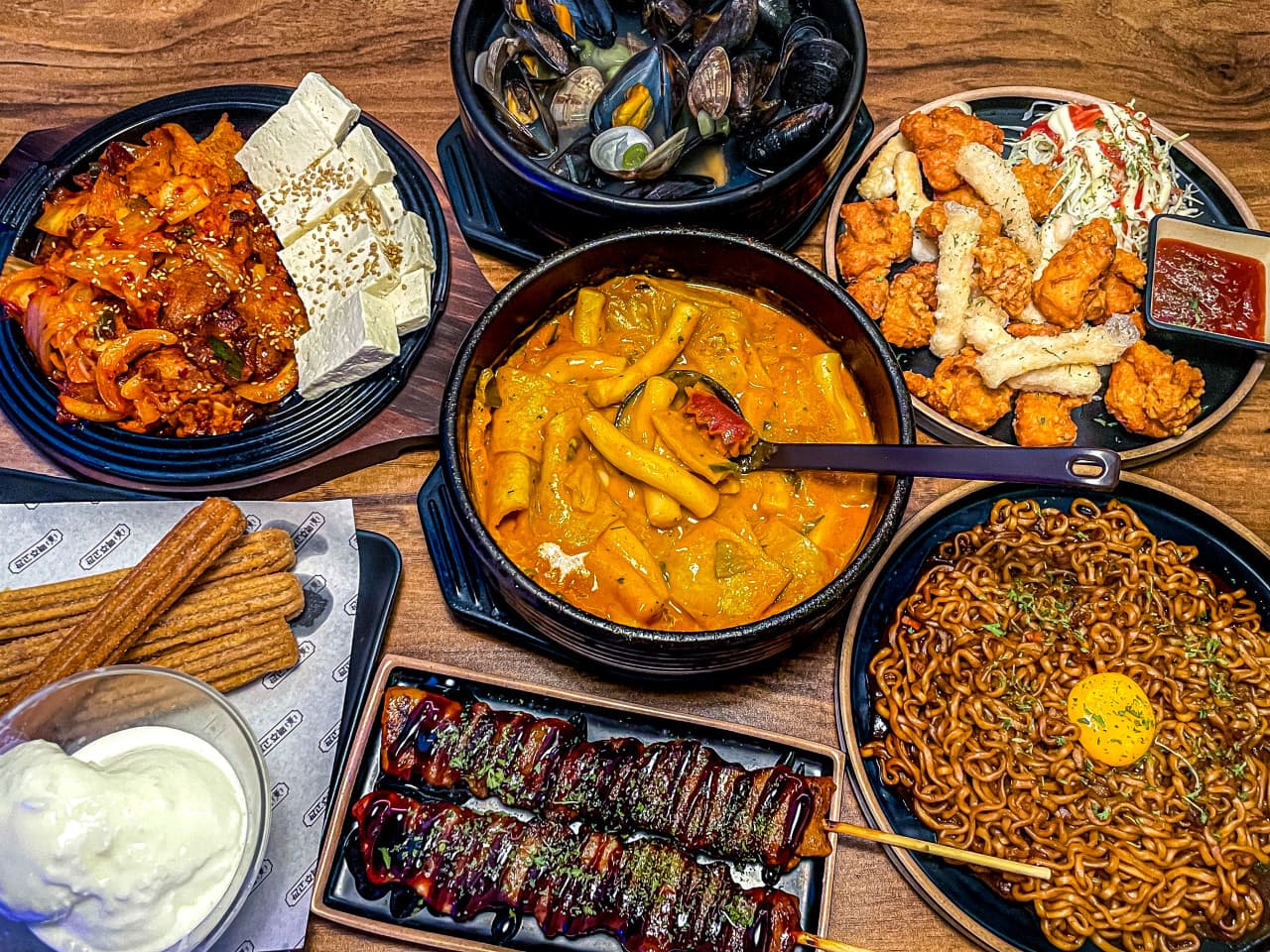 나무 테이블 위 떡볶이 뚝배기와 닭꼬치, 계란 노른자를 올린 짜장 라면 등 여러 접시에 나뉜 안주 구성