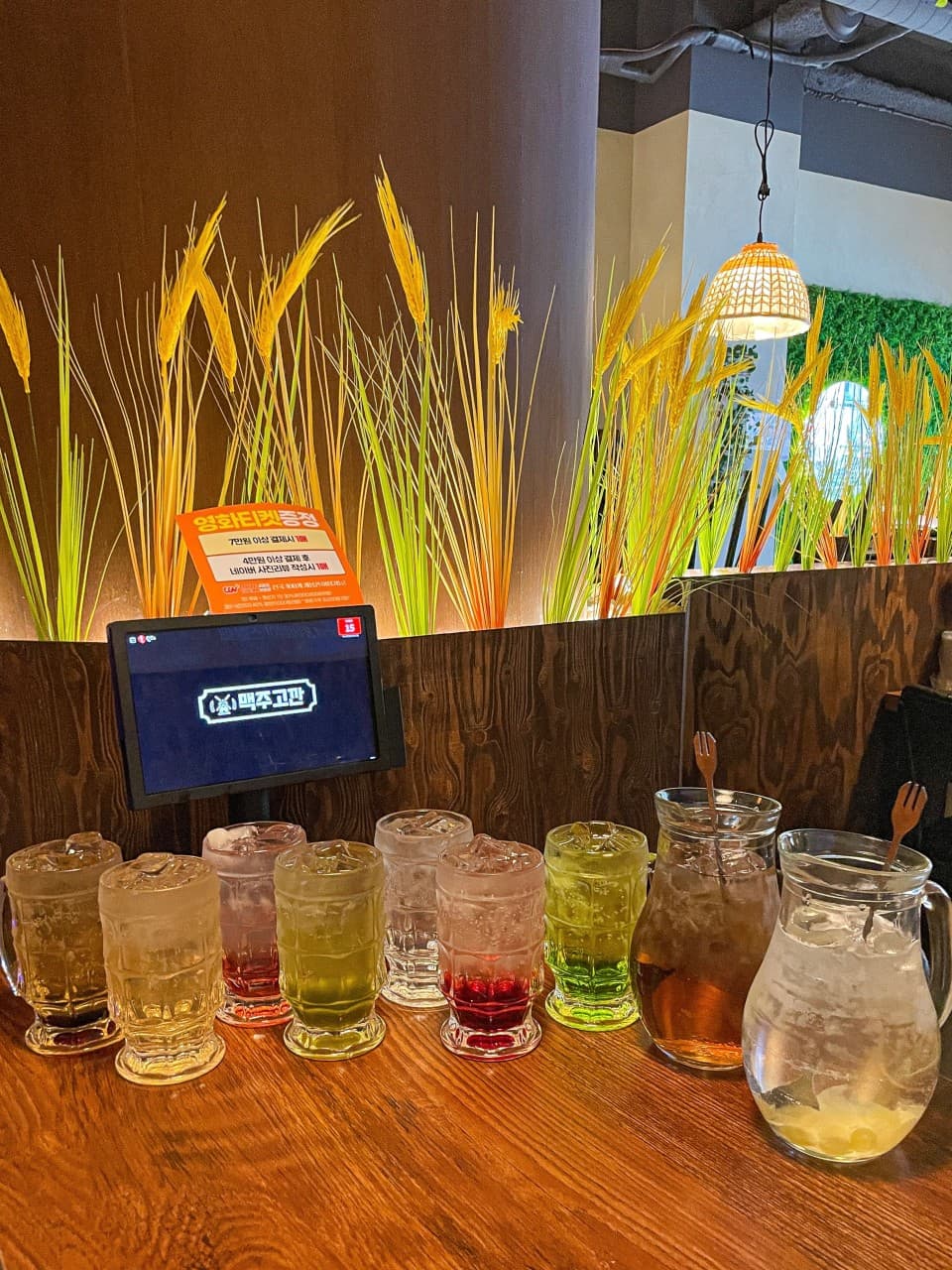 나무 테이블 위 얼음이 담긴 다양한 색의 하이볼 잔과 투명 피처. 보리 조형물로 장식된 칸막이와 개별 주문용 태블릿이 있는 좌석 전경