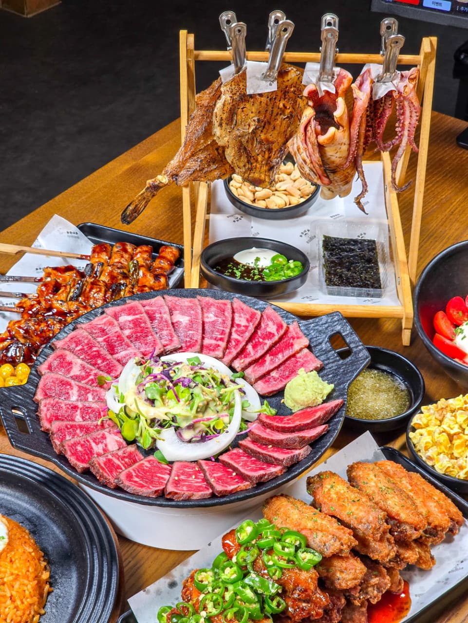 둥글게 배치된 소고기 타다끼와 닭날개 튀김, 닭꼬치, 나무 스탠드에 매달린 마른안주가 차려진 1인용 목재 테이블 전경