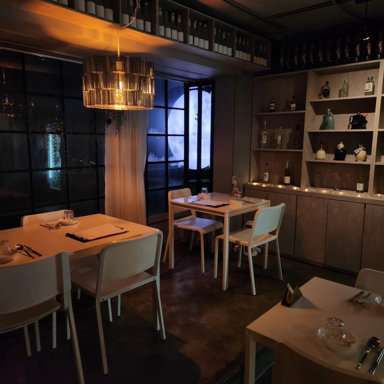 주황색 조명 아래 놓인 흰색 테이블과 의자. 창밖의 둥근 달 조형물과 벽면 선반에 진열된 술병들이 보이는 어두운 실내 전경.