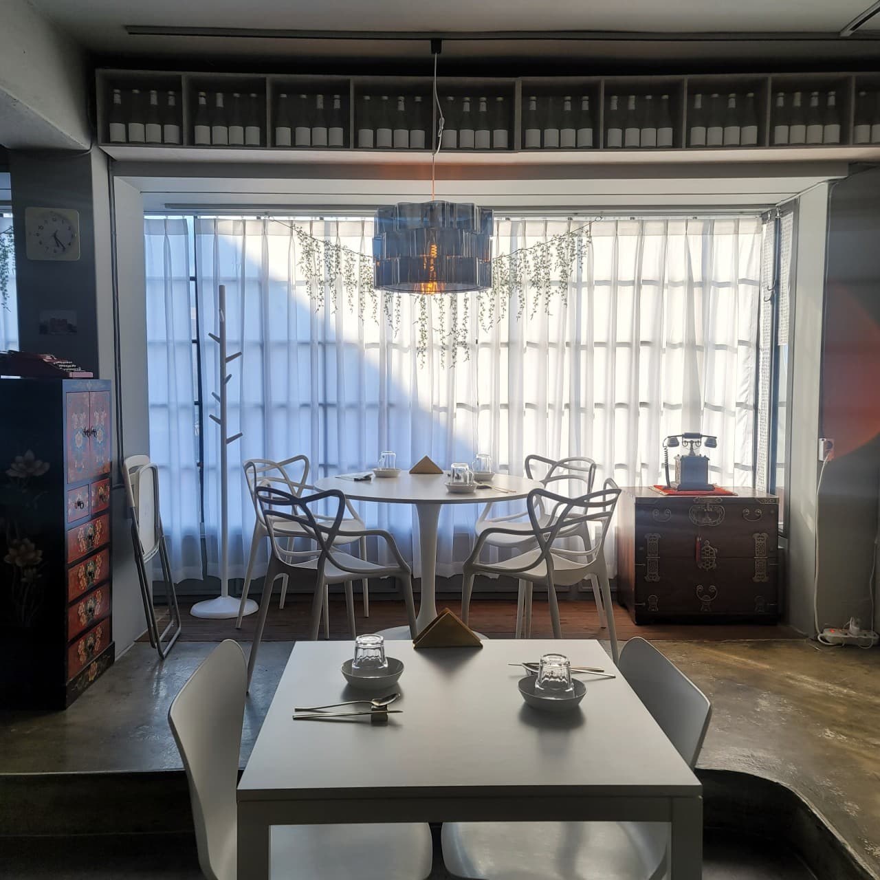 흰색 커튼 앞 원형 테이블과 1인용 식기가 놓인 사각 식탁. 벽면 상단 선반의 술병들과 전통 목제 가구가 배치된 내부 공간.