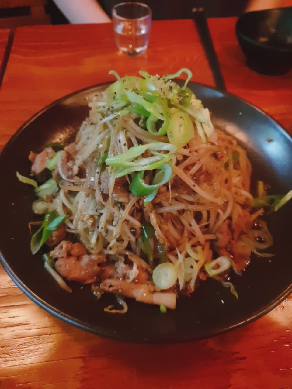 붉은 조명이 비치는 목재 테이블 위 숙주 고기 볶음과 대파 고명이 담긴 검은 접시와 작은 술잔