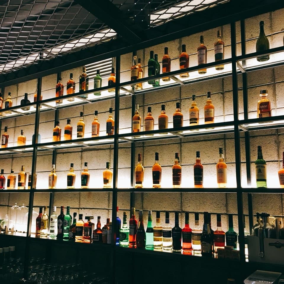 검은색 금속 선반에 정렬된 위스키 병들과 하단 조명이 비치는 콘크리트 벽면. 바 카운터 좌석에서 바라본 술병 진열장 전경