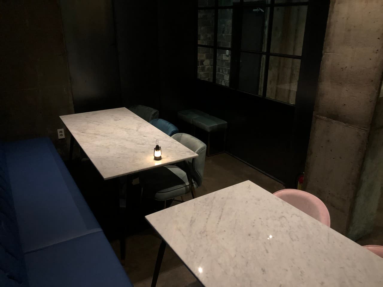 어두운 실내 공간에 배치된 흰색 대리석 테이블과 작은 램프, 벽면을 따라 놓인 긴 소파와 개별 의자