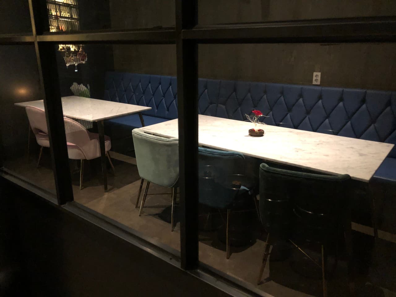 창틀 너머로 보이는 파란색 퀼팅 벽면 소파와 대리석 테이블. 분홍색과 청록색 1인용 의자가 배치된 어두운 조명의 바 내부 전경