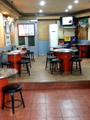 붉은 원통형 테이블과 검은색 스툴이 배치된 실내. 단차가 있는 바닥에 놓인 원형 좌석들과 벽면의 화이트보드 메뉴판, TV, 적재된 주류 상자가 보이는 전경