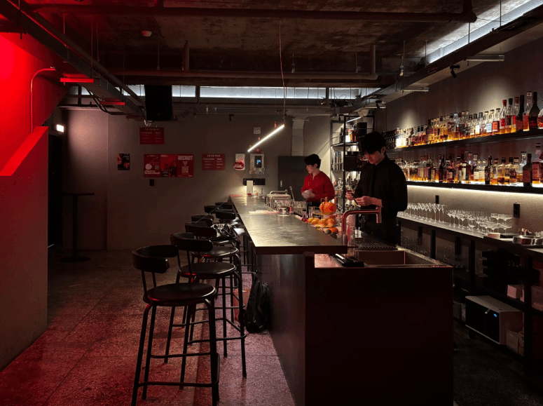 부산광역시 동구 혼술 맥주 호프 체리베이스먼트 - 붉은 조명 아래 긴 다찌석과 벽면 선반의 다양한 술병들이 진열된 1인석 바 테이블 공간