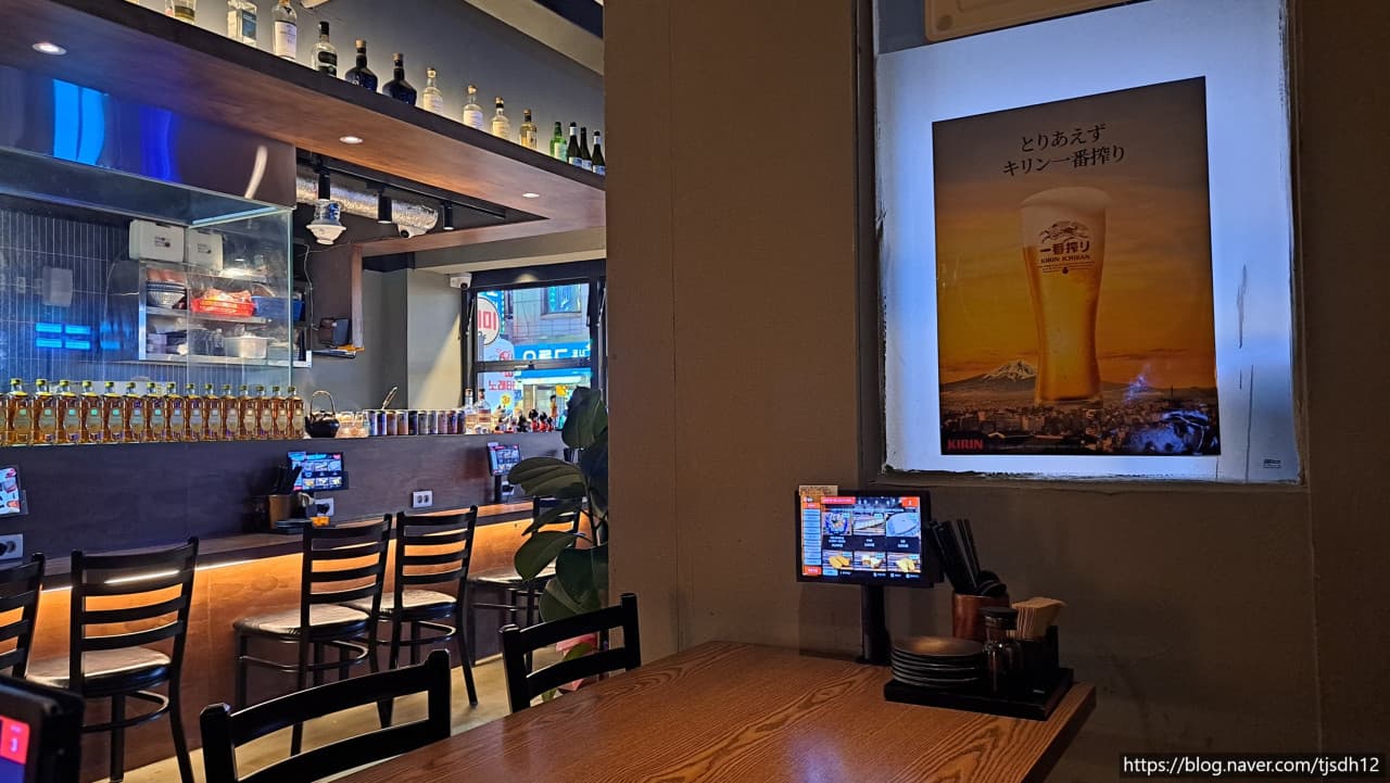 부산광역시 중구 히노카오뎅 남포점 요리주점. 주황색 조명이 비추는 혼술 다찌석과 위스키 병이 나열된 선반, 벽면의 맥주 포스터와 테이블 키오스크 전경.