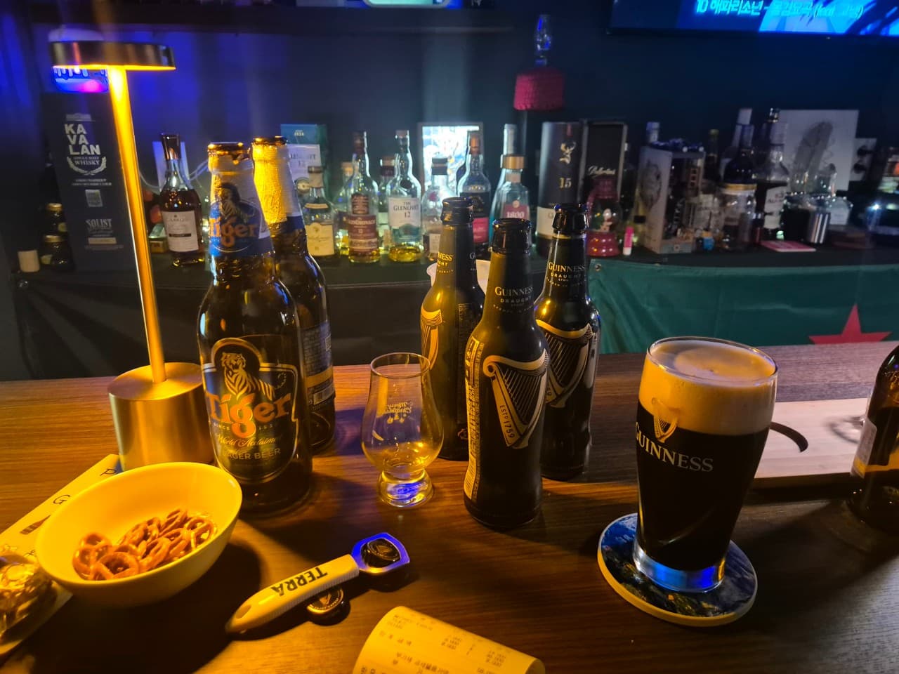 부산광역시 중구 펍송 인 광복 맥주 호프 - 노란 램프 조명이 비추는 다찌석 위 기네스와 타이거 맥주병. 거품이 올라온 흑맥주와 프레첼이 놓인 1인 혼술 상황.