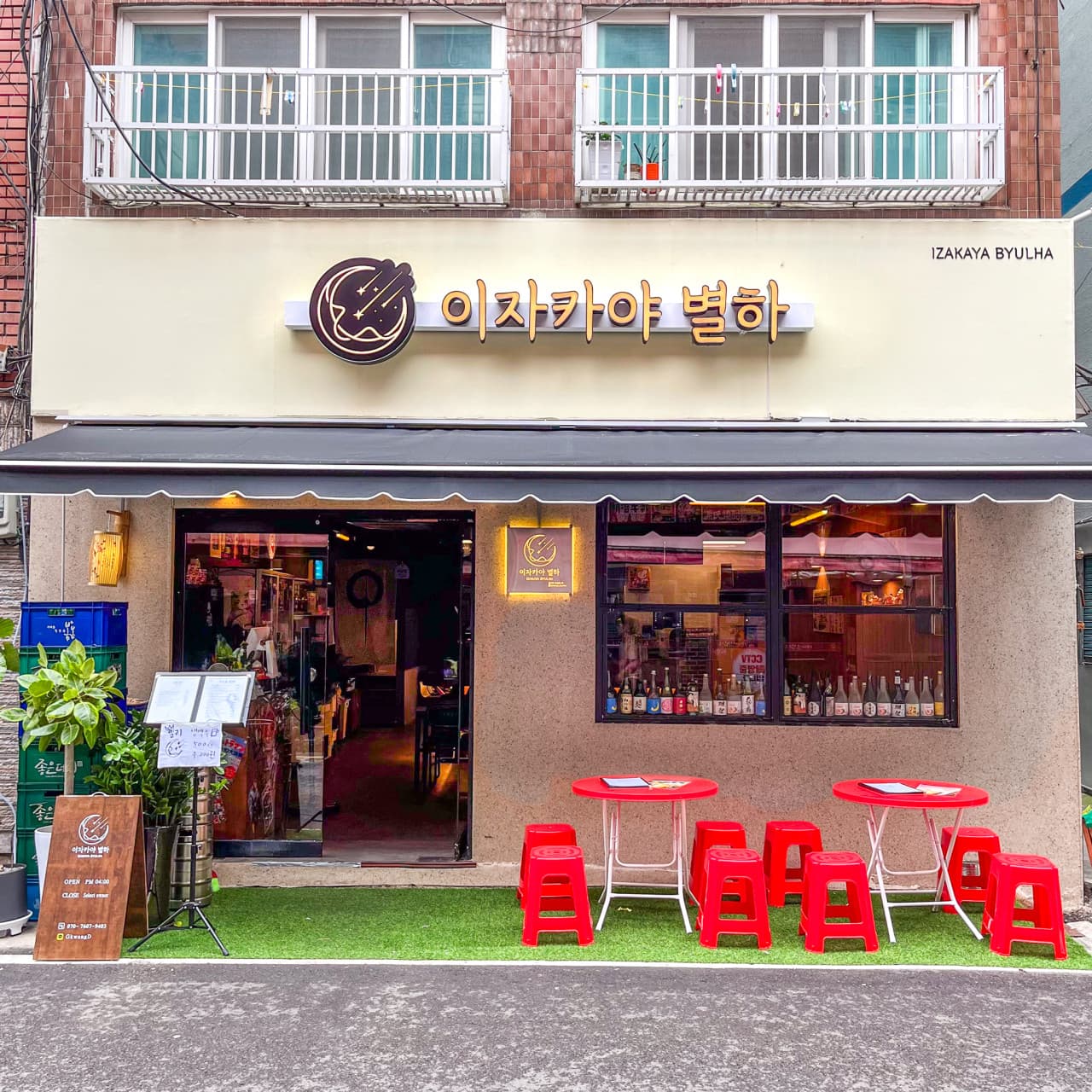 부산광역시 중구 이자카야별하 입구 앞 빨간색 야외 테이블과 인조잔디. 주황색 조명이 켜진 실내 다찌석과 창가에 진열된 사케 병이 보이는 1인 혼술 이자카야 전경.