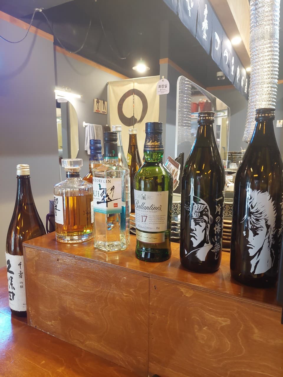 부산광역시 중구 혼술 이자카야별하 - 나무 다찌석 위 위스키와 사케 병들, 조명 아래 1인 바 테이블이 배치된 이자카야 내부 풍경