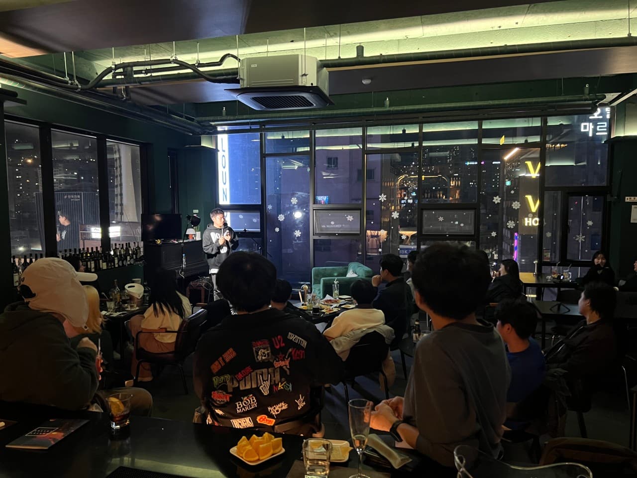 부산광역시 중구 선셋라운지 바 - 통창 너머 도시 야경이 보이는 실내 조명 아래 1인 다찌석에 놓인 과일과 술. 공연을 관람하며 혼술하는 상황.