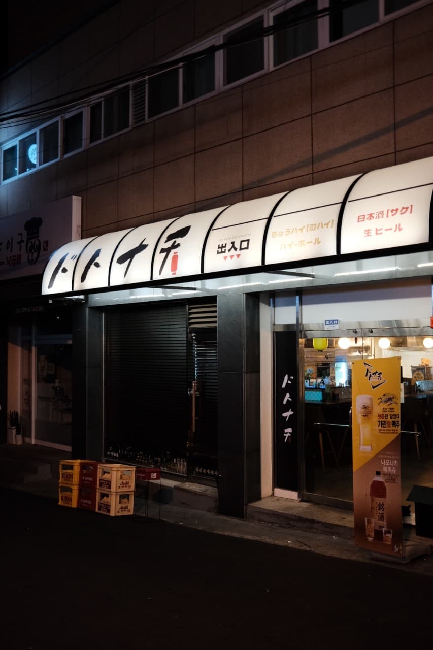 경상남도 창원시 이자카야 도토나치 창원 - 하얀 조명 간판 아래 1인 다찌석이 보이는 매장 입구와 노란 맥주 박스가 놓인 혼술 공간