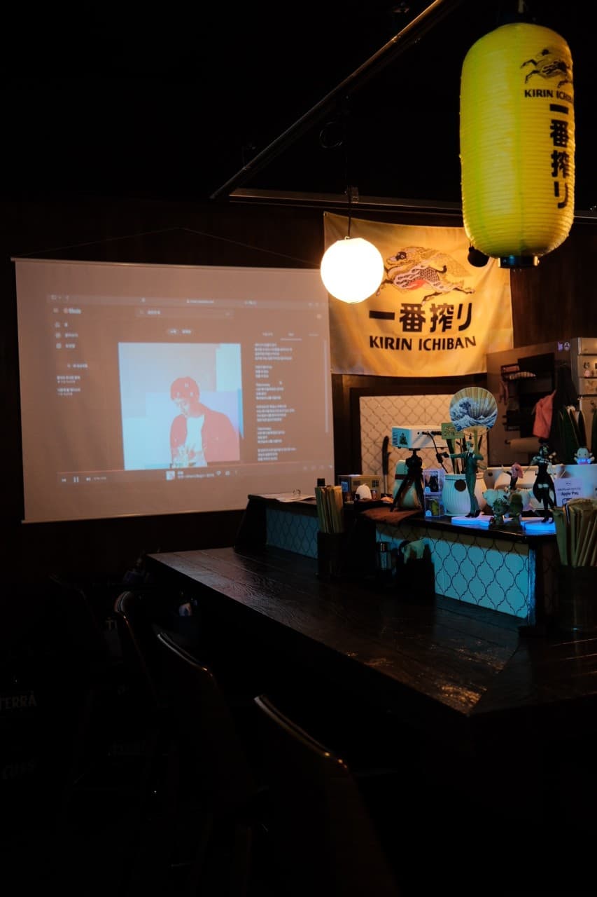 경상남도 창원시 이자카야 도토나치 창원 노란 제등과 빔프로젝터가 보이는 1인 다찌석. 어두운 배경 속 조명 아래 목재 바 테이블이 배치된 혼술 공간.