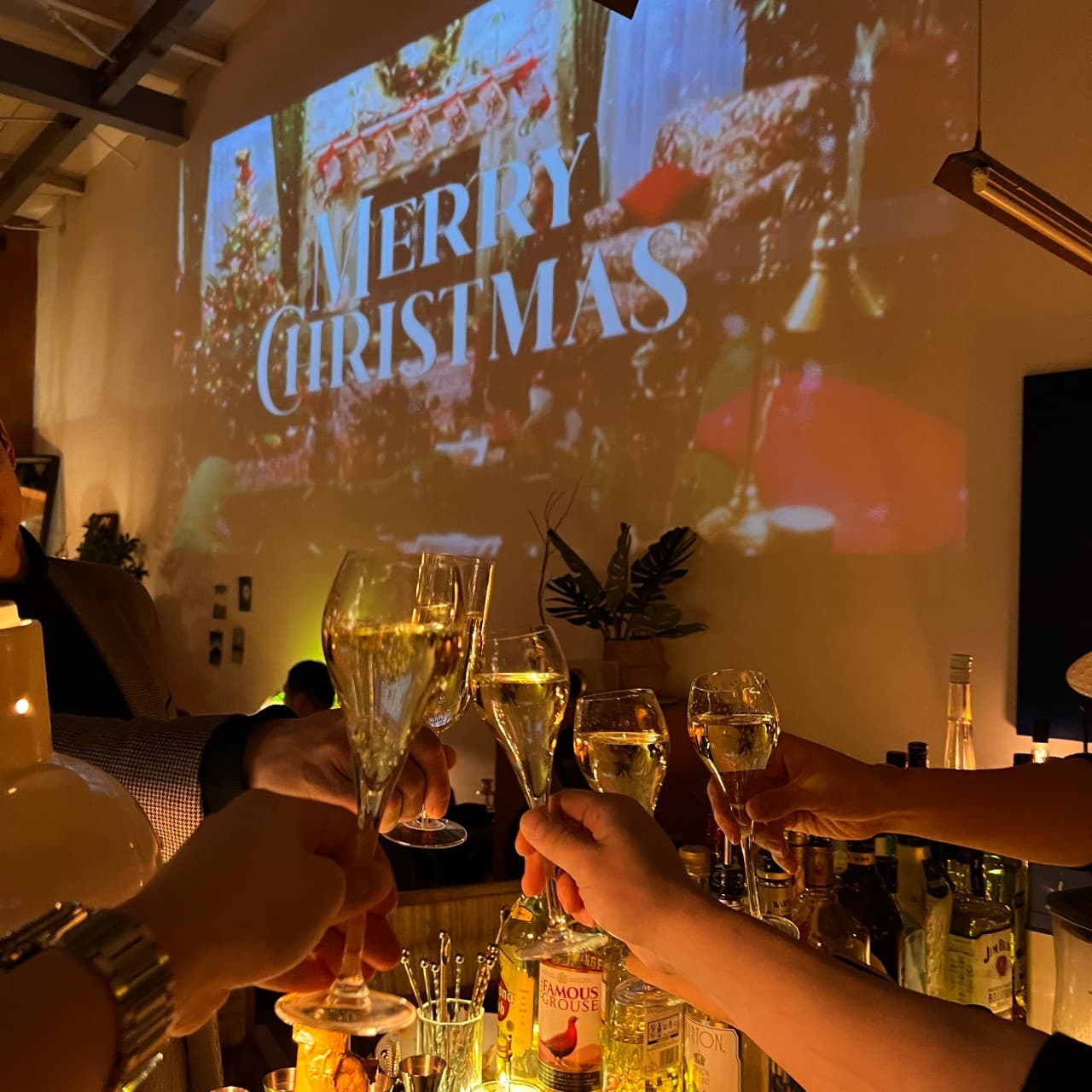 경상남도 사천시 진공관 다찌석의 혼술 분위기. 벽면 메리 크리스마스 빔 영상 아래 술잔을 부딪히는 상황과 바 카운터에 진열된 위스키 병들이 보이는 기타 주점.