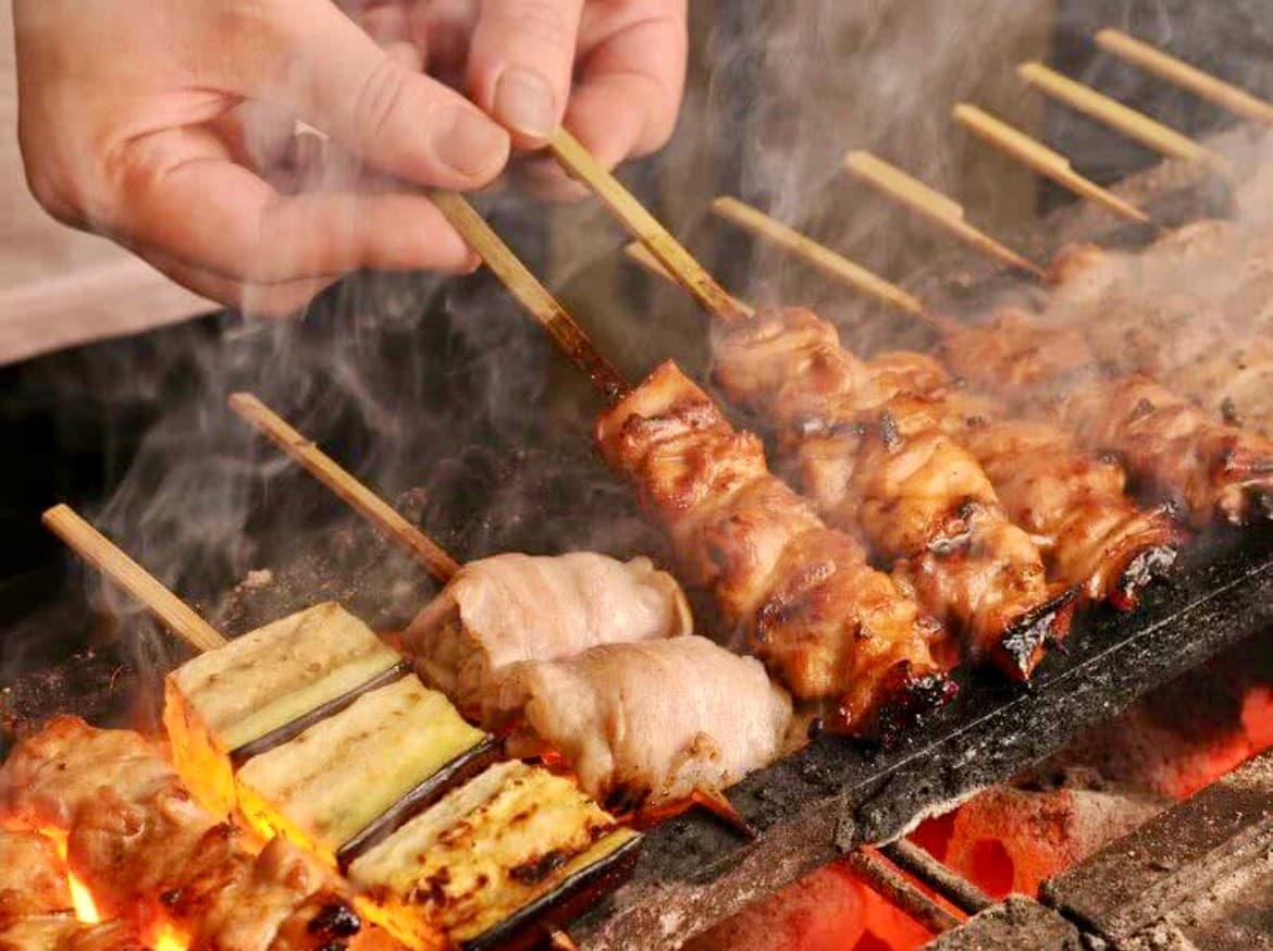 경상남도 사천시 혼술 이자카야 야키토리 낙원 - 숯불 빛 아래 연기가 피어오르는 꼬치구이를 뒤집는 손과 다찌석 1인 메뉴 조리 상황