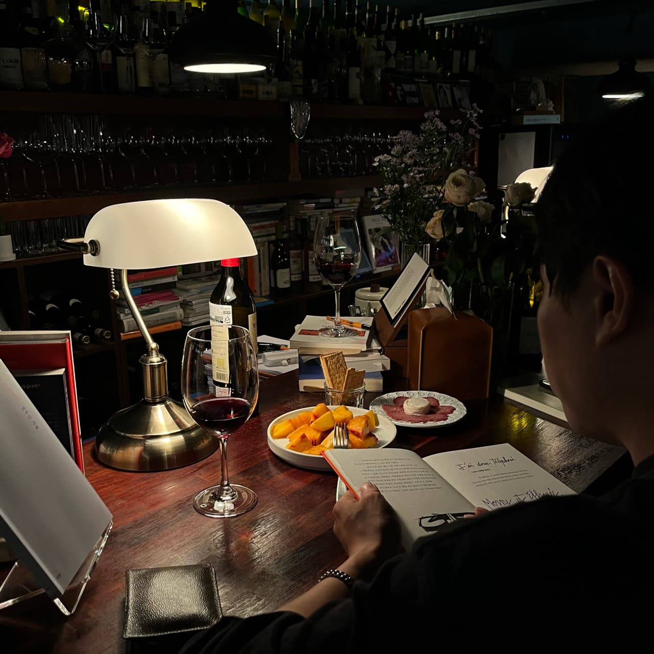 경상남도 진주시 Bar 사건의장소 다찌석에서 스탠드 조명 아래 레드 와인과 과일을 곁들여 책을 읽는 1인 혼술. 목재 테이블 위 와인잔과 안주가 놓인 상황.
