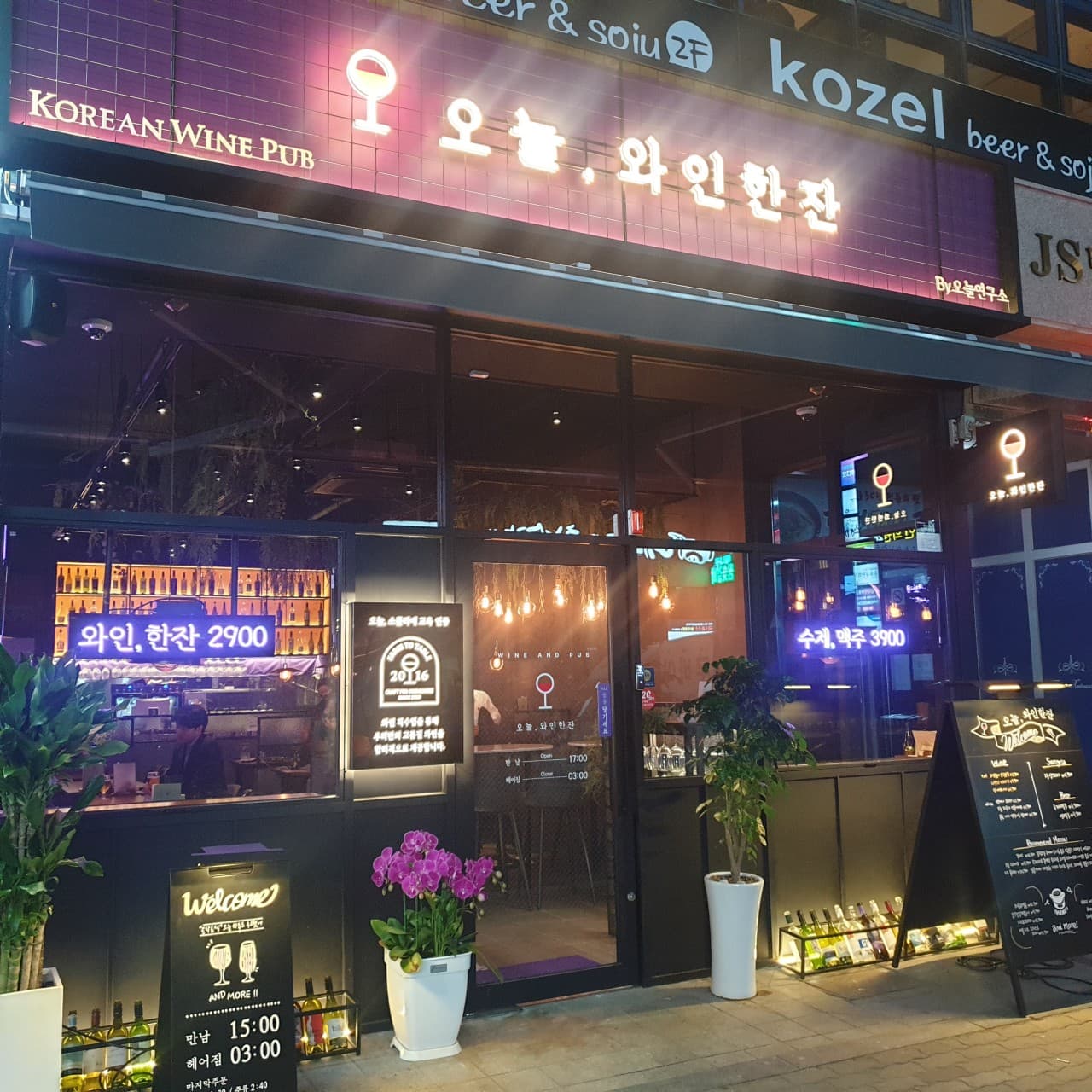 경상남도 진주시 오늘와인한잔 진주혁신점 입구. 보라색 네온 조명이 켜진 유리창 너머로 혼술 전용 다찌석과 와인 진열대, 메뉴판이 배치된 모습.