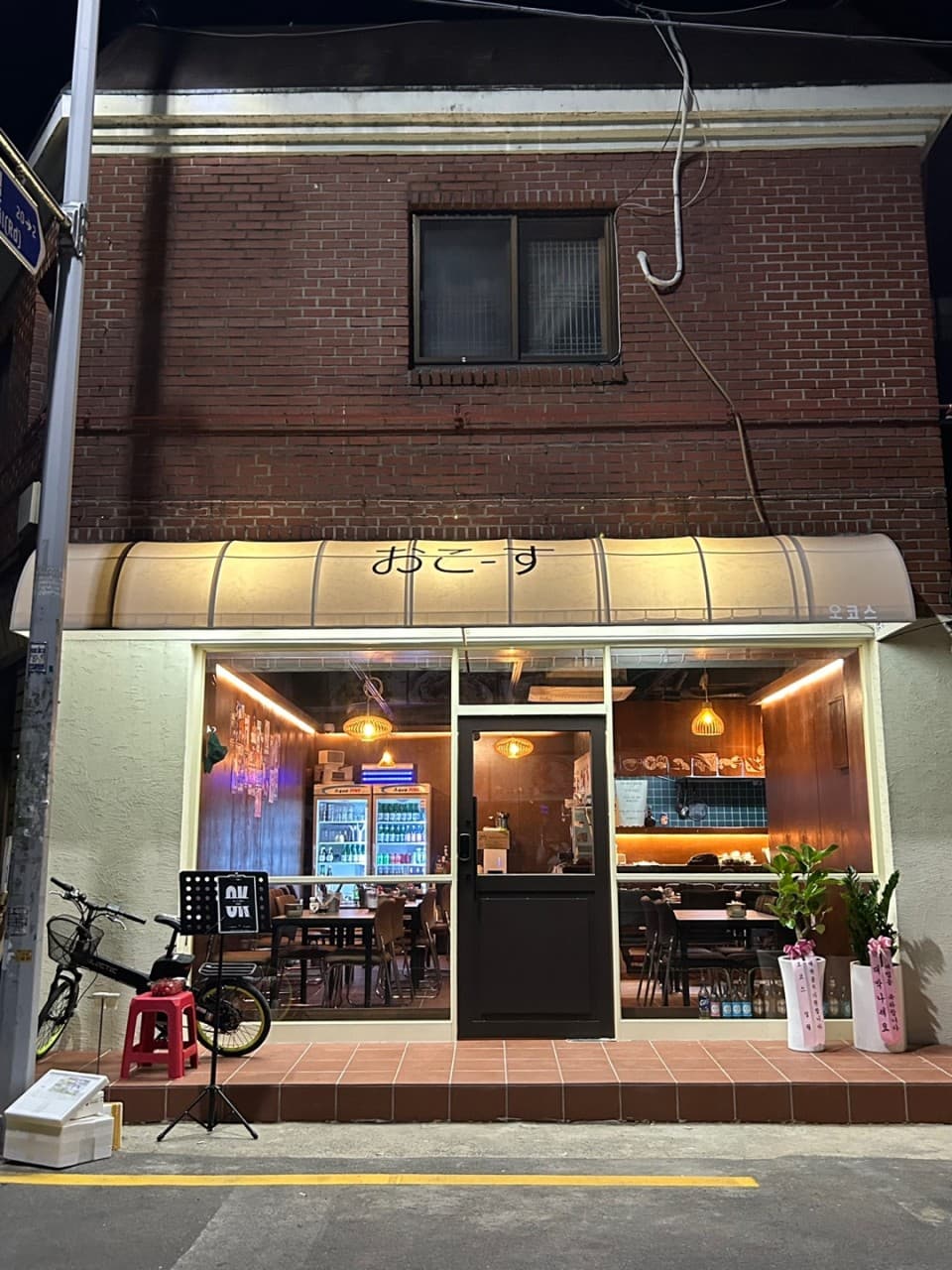 경상남도 진주시 요리주점 오코스 진주칠암본점 진주. 주황색 조명 아래 다찌석과 1인 좌석이 배치된 통유리창 내부와 매장 앞 자전거가 놓인 밤거리 혼술 상황 묘사.