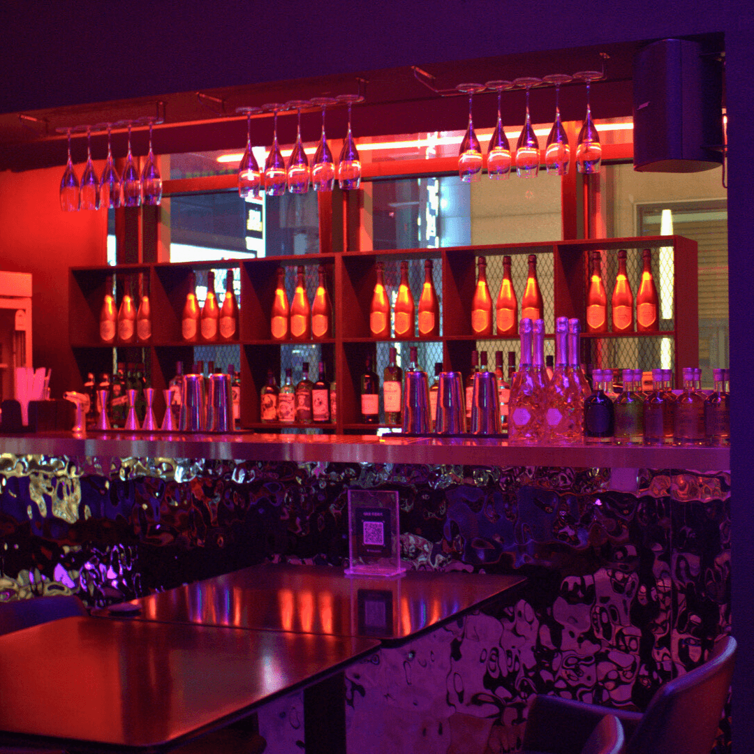 서울특별시 송파구 나쉬펍 잠실방이점 맥주 호프의 붉은 네온 조명 아래 물결무늬 바 테이블과 다찌석. 진열된 술병과 와인잔이 비치는 공간에서 즐기는 1인 혼술 상황.
