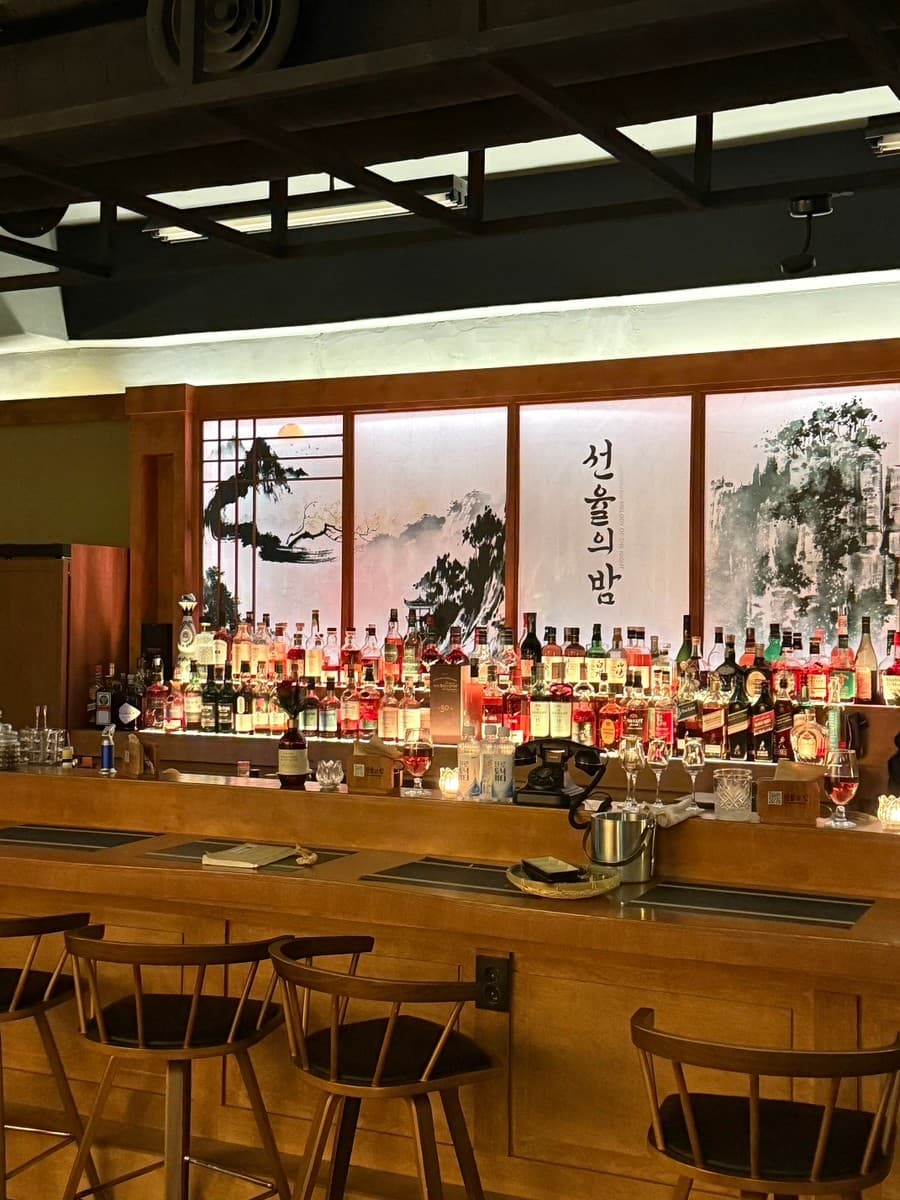 서울특별시 송파구 선율의밤 잠실방이점 바(Bar). 동양적인 수묵화 배경과 조명 아래 진열된 다양한 위스키병, 목재 소재의 긴 다찌석과 1인 좌석이 마련된 혼술 상황.