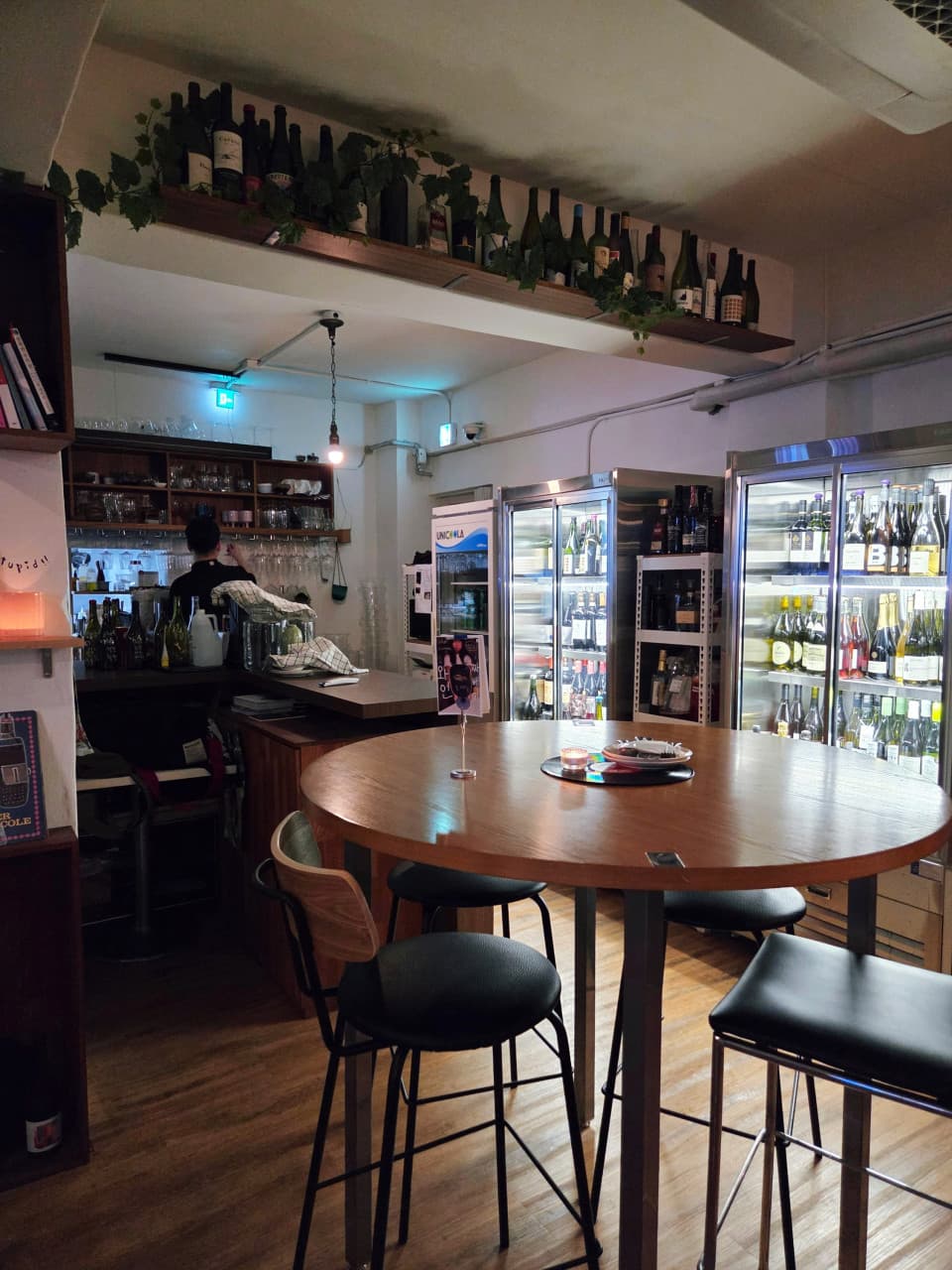 서울특별시 송파구 요리주점 디에스티클럽 석촌 내부. 목재 원형 테이블과 1인 다찌석, 와인 냉장고가 배치된 혼술 공간. 주황빛 핀조명이 술병과 잔을 비추는 실내 구성.