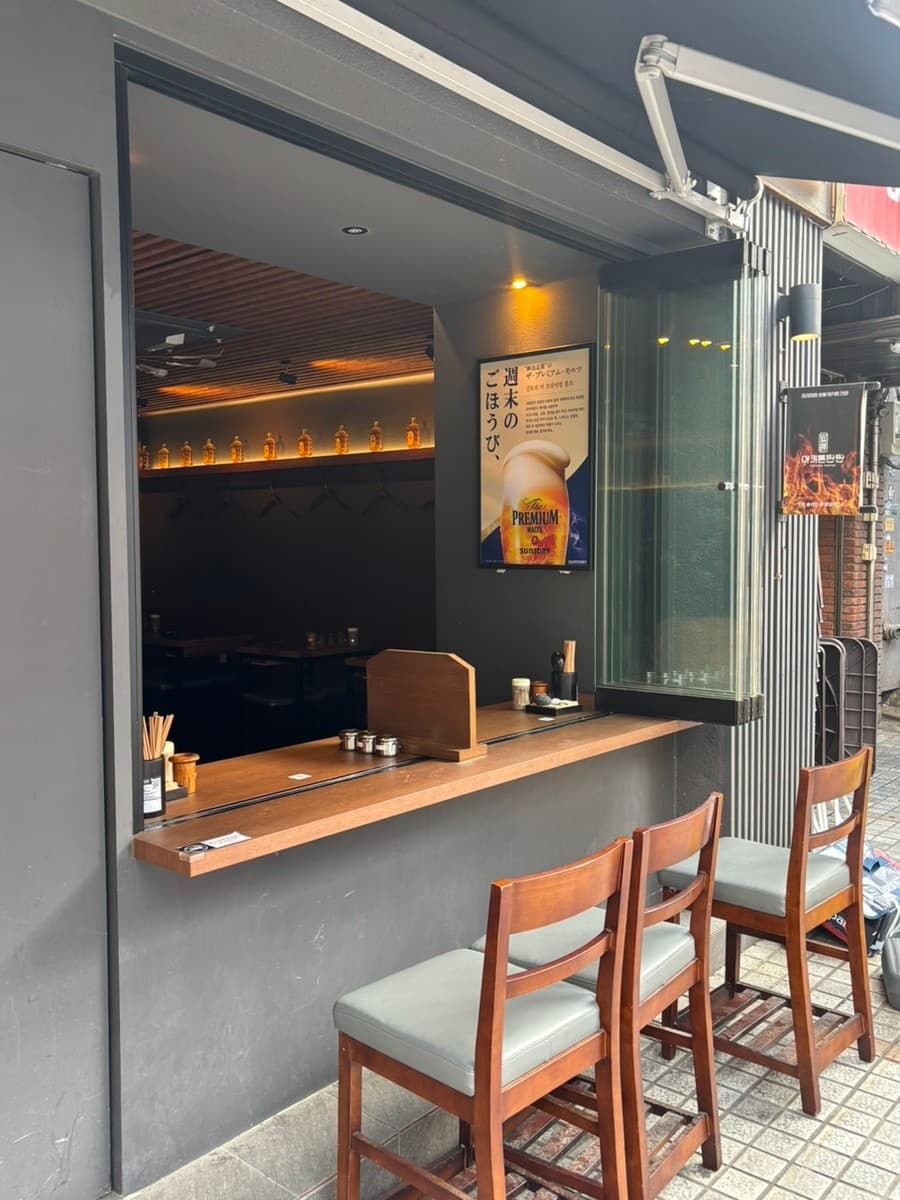 서울특별시 송파구 야키톤탄탄 잠실 이자카야의 외부 테라스 다찌석. 나무 소재 1인 좌석과 노란 조명이 비치는 실내 선반 위 술병들이 보이는 혼술 상황의 창가 카운터 테이블.