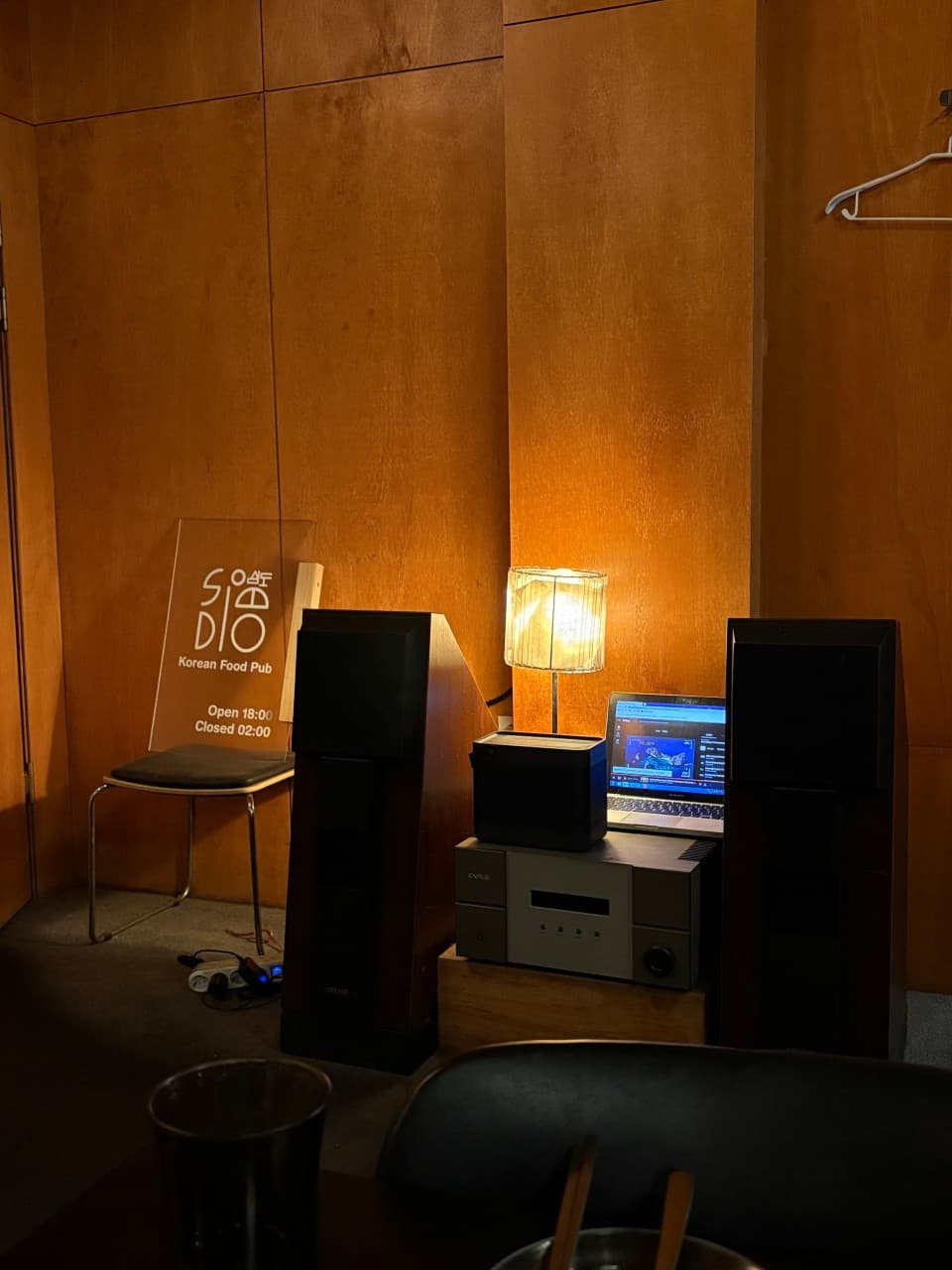 대전광역시 서구 요리주점 심도 대전서구 - 주황색 조명과 나무 벽면, 대형 스피커가 배치된 다찌석에서 즐기는 1인 혼술 상황. 스탠드 조명 아래 오디오 장비와 좌석이 위치함.