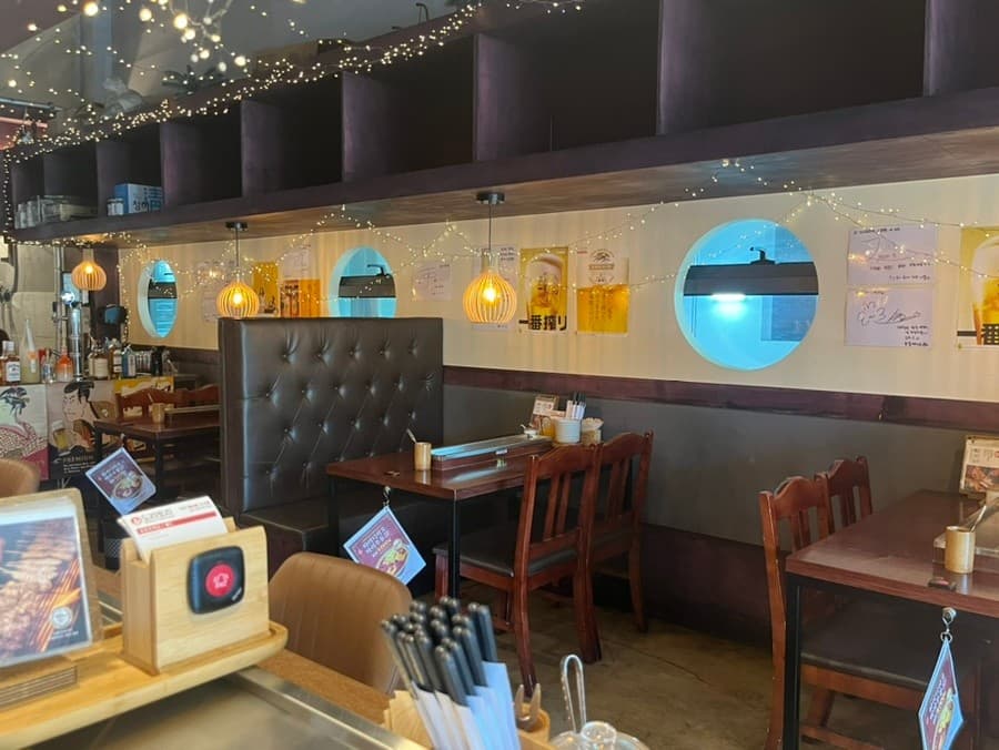 대전광역시 유성구 도리토리 대전봉명점 요리주점 - 전구 조명 아래 1인 다찌석과 목재 테이블. 원형 창문이 있는 벽면과 정돈된 혼술 공간.