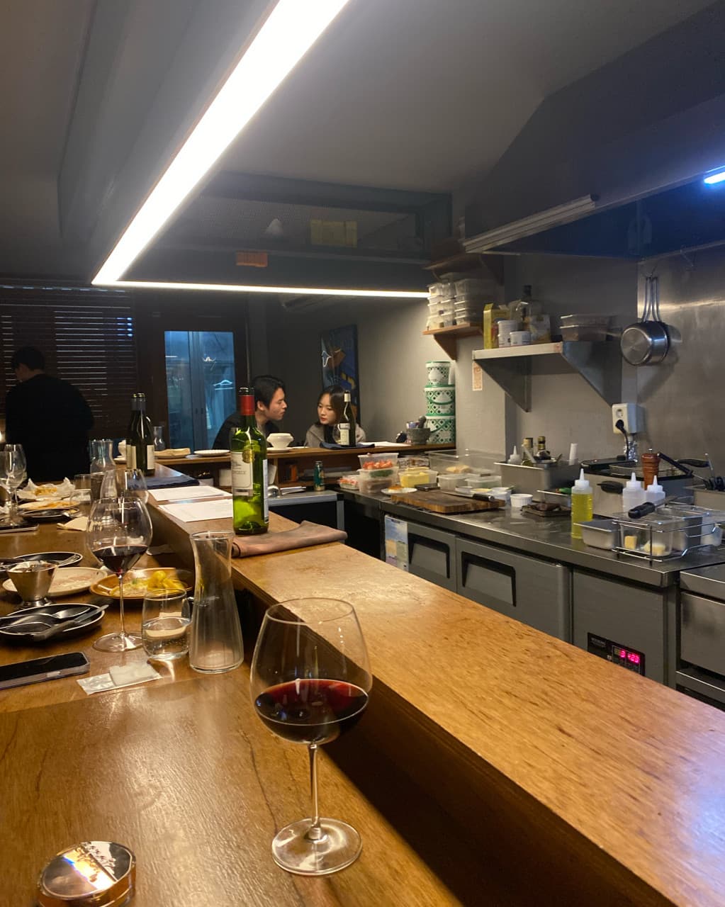 대구광역시 중구 와인바 러프 대구 동성로 - 직선형 조명이 비추는 목재 다찌석 위 레드 와인 한 잔과 오픈 키친 앞 1인 혼술 상황